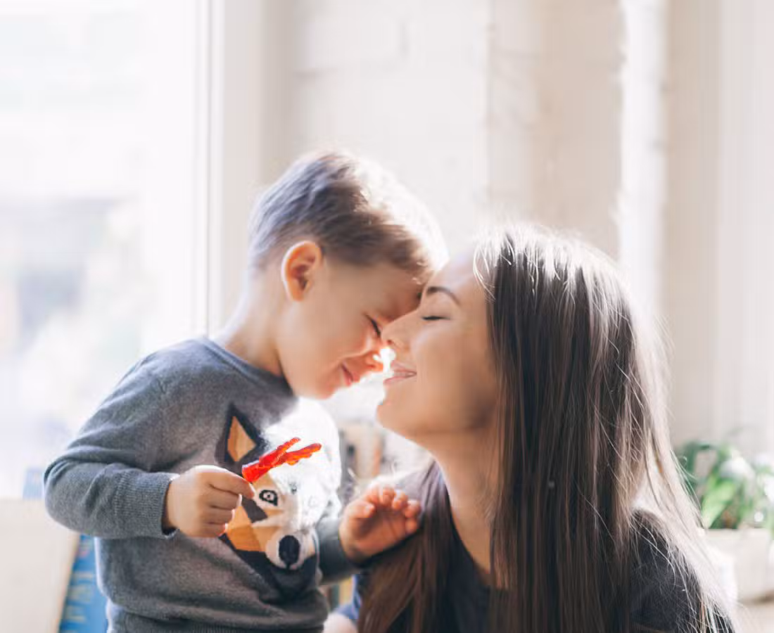 A young child and a woman smile with their faces touching gently in a sunlit room. The child wears a gray shirt with an animal graphic, and both appear joyful, sharing a tender and happy moment together.