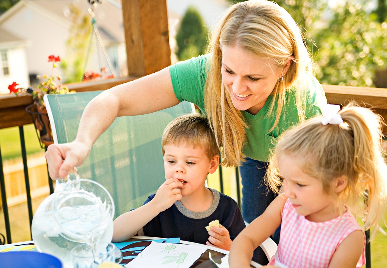 A woman in a green shirt pours water from a glass pitcher for two young children sitting at an outdoor table. The boy is eating a snack, and the girl in a pink dress is drawing. It is sunny and they are on a porch.