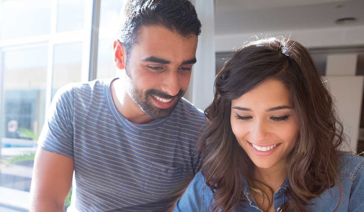 A smiling couple sits on a couch at home, looking at a smartphone together. The background shows a bright, cozy kitchen with shelves, plants, and kitchenware.