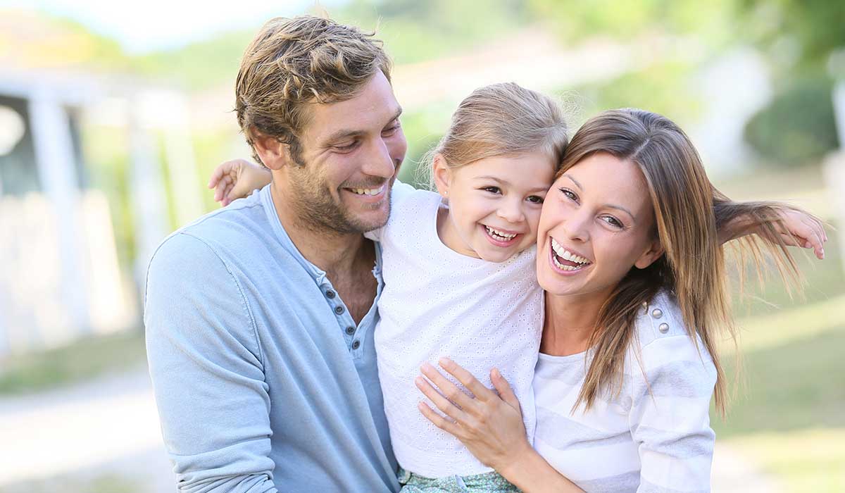 A happy family of three‚Äîan adult man, an adult woman, and a young girl‚Äîsmiles and embraces each other outdoors on a sunny day. The girl is between the adults, who both look joyful and relaxed.