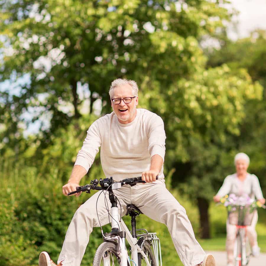 An older man in light-colored clothes rides a bicycle joyfully with his legs outstretched, while an older woman follows behind him on a bike; both are outdoors on a sunny day with greenery in the background.