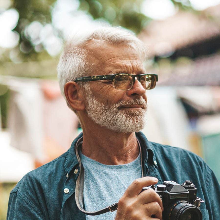 An older man with gray hair and a beard, wearing glasses and a blue shirt, holds a vintage camera outdoors. The background is blurred with greenery and sunlight visible.