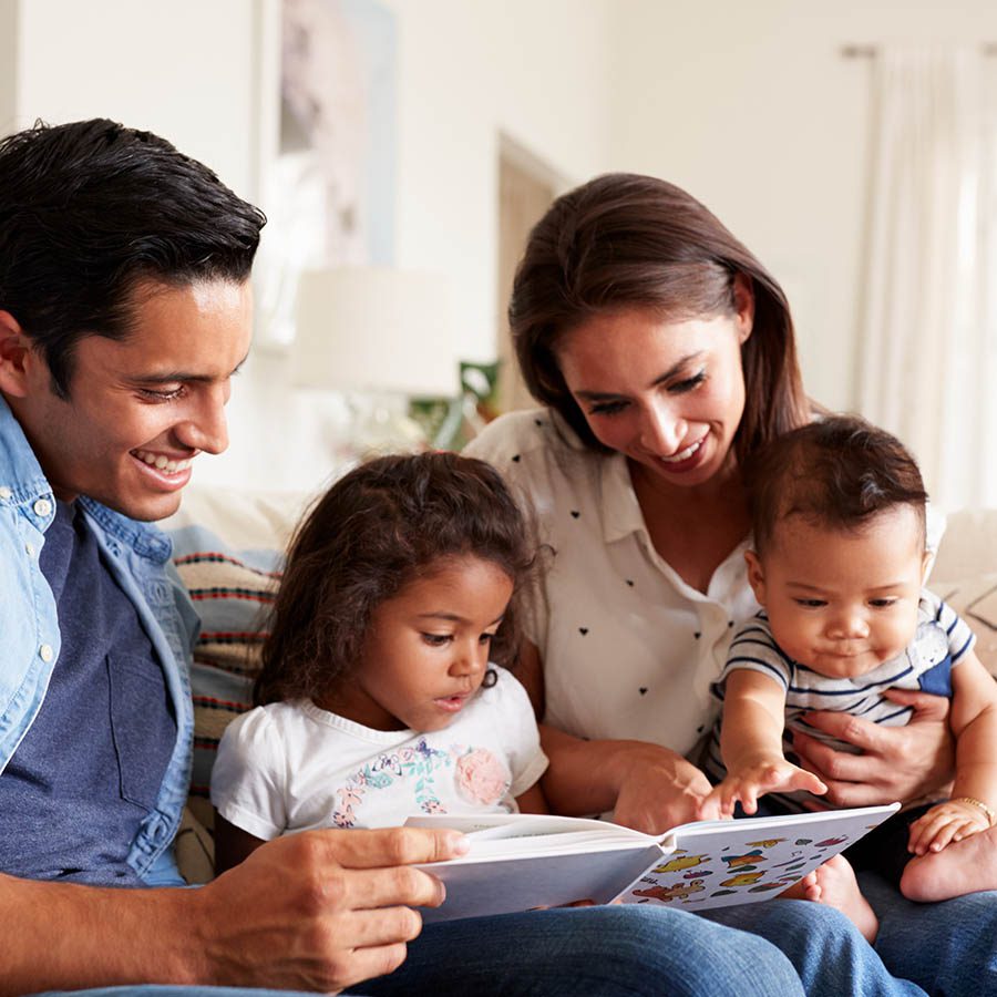 A family of four sits on a couch reading a book together. The parents smile as their young daughter and baby look at the pages, enjoying a cozy and happy moment in a bright living room.