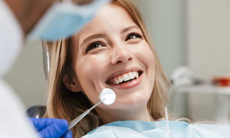 A woman sits in a dental chair, smiling while a dentist wearing gloves and a mask holds a dental mirror near her mouth during a dental checkup.